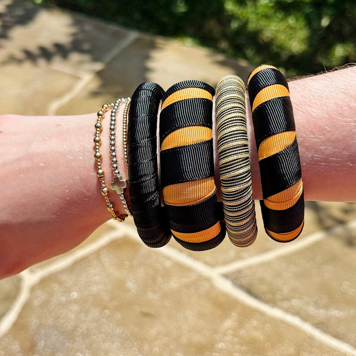 Close-up of a wrist wearing multiple black and gold Butter Republic bracelets on a stone surface with greenery in the background