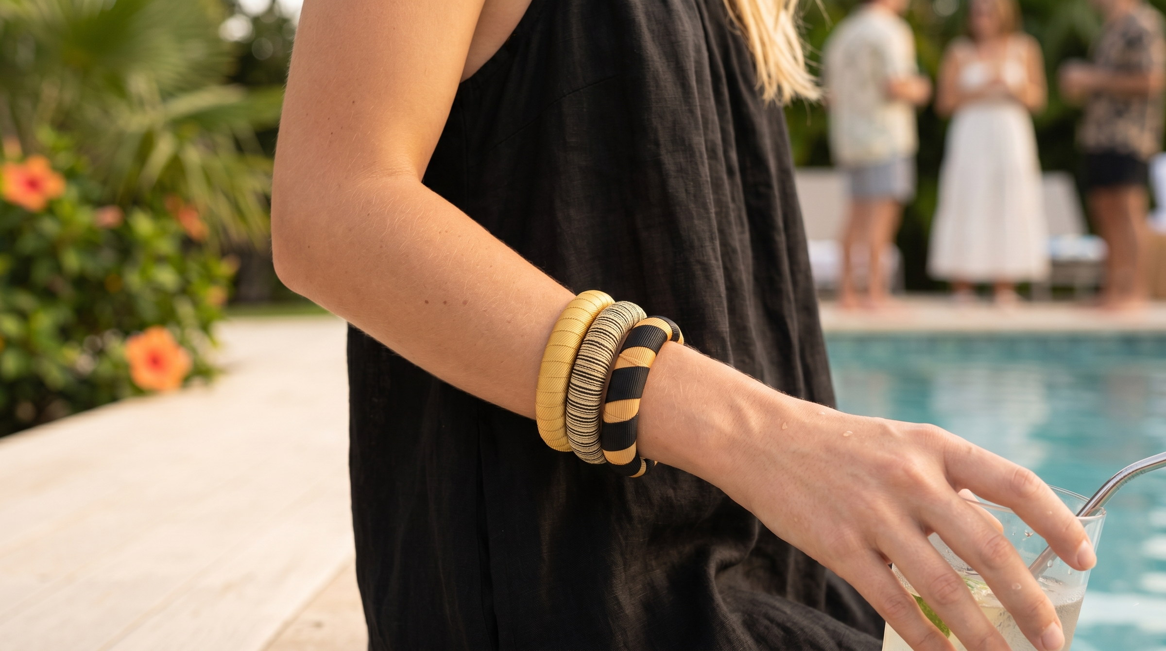 woman wearing three gold and black Butter Republic bracelets; standing outdoors near a swimming pool with people blurry in the background