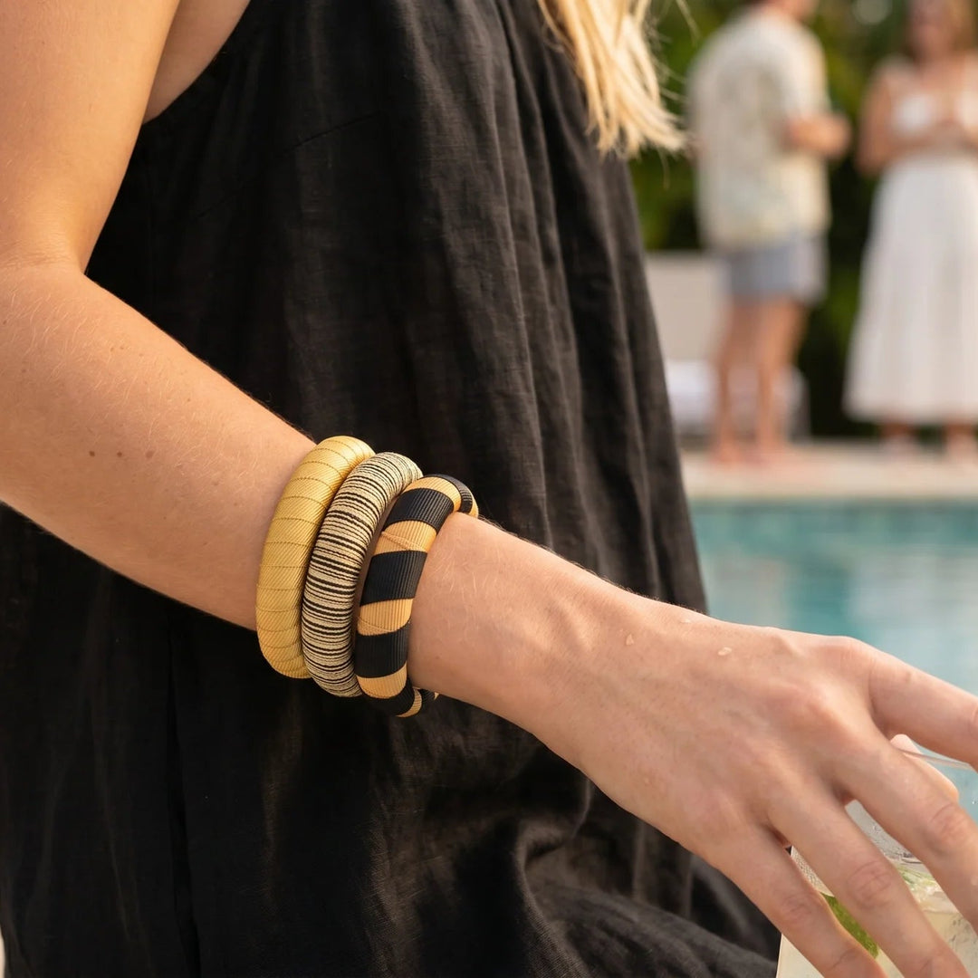 Woman by a pool wearing a black sleeveless top and a stack of black and gold Butter Republic bracelets, with blurred people and plants in the background.