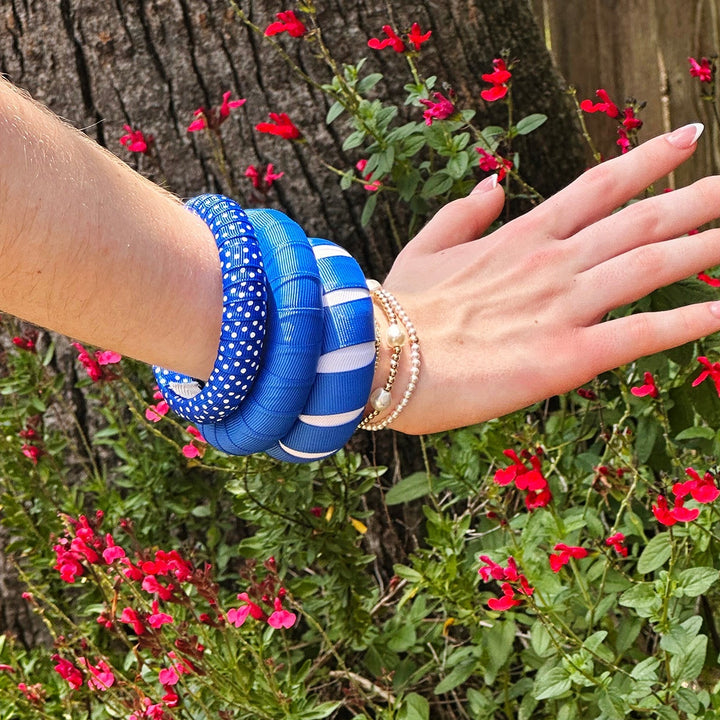 Hand wearing a stck of Butter Republic blue and white bracelets with flowers in the background