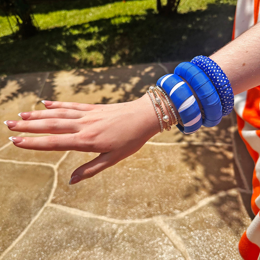 Hand wearing multiple blue and white Butter Republic bracelets on a stone surface with grass and wooden fence in the background