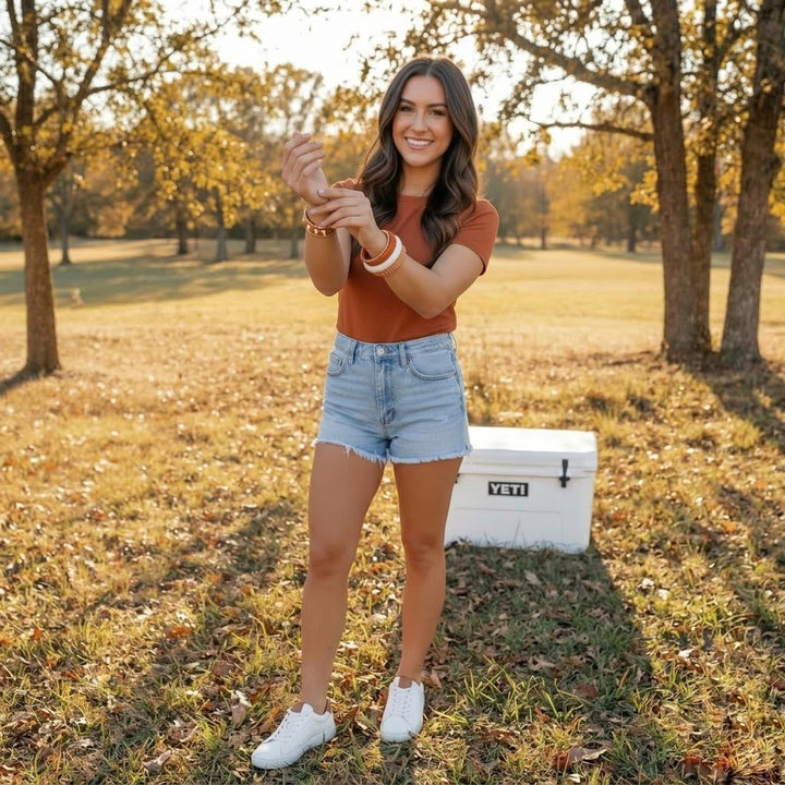woman in a field, wearing burnt orange Butter Republic bracelets, with a cooler in the background