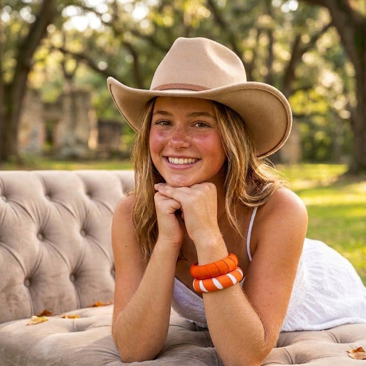 young woman wearing orange and white Butter Republic bracelets and a beige cowboy hat, leaning over an ottoman outdoors