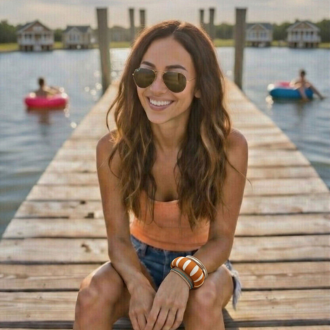 woman sitting on a wooden dock by a lake, wearing a burnt orange and white Butter Republic bracelet, aviator sunglasses, and a burnt orange top