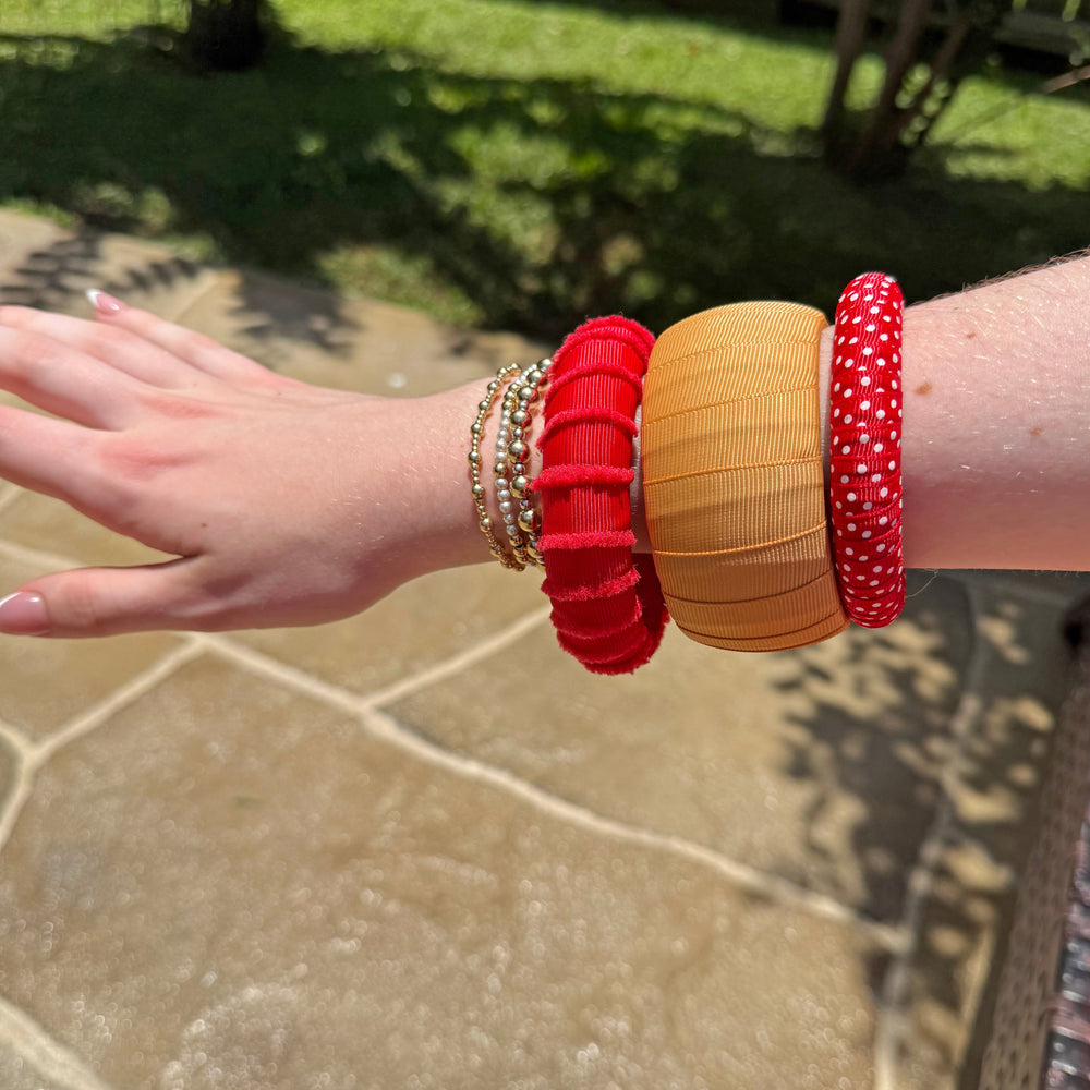 Woman wearing multiple red and gold Butter Republic bracelets on a stone surface with grass and wooden fence in the background