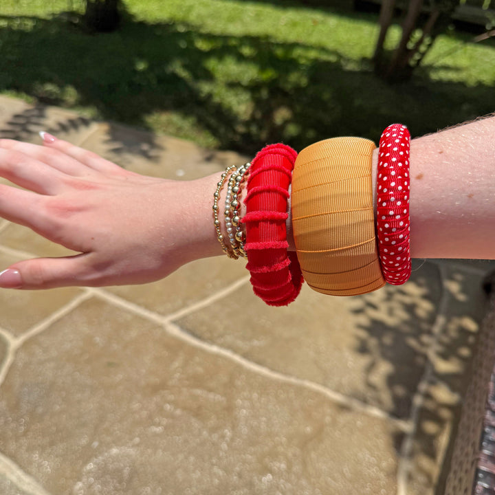 Woman wearing multiple red and gold Butter Republic bracelets on a stone surface with grass and wooden fence in the background
