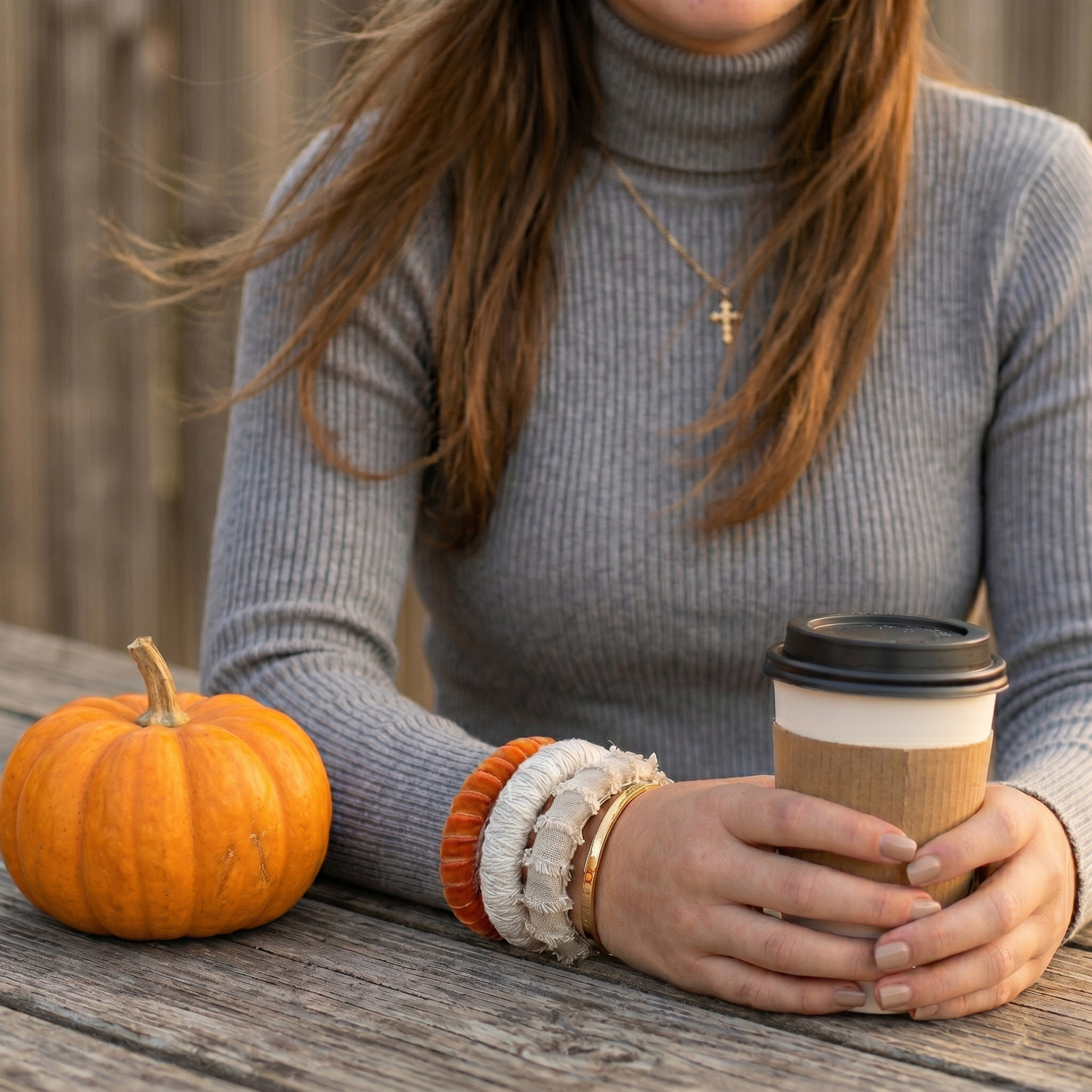 woman wearing a Butter Republic burnt orange velvet bracelet and two neutral colored Butter Republic bracelets and a ribbed gray turtle neck top, sitting at a wooden table outdoors, holding a coffee cup with a small orange pumpkin on the table next to her