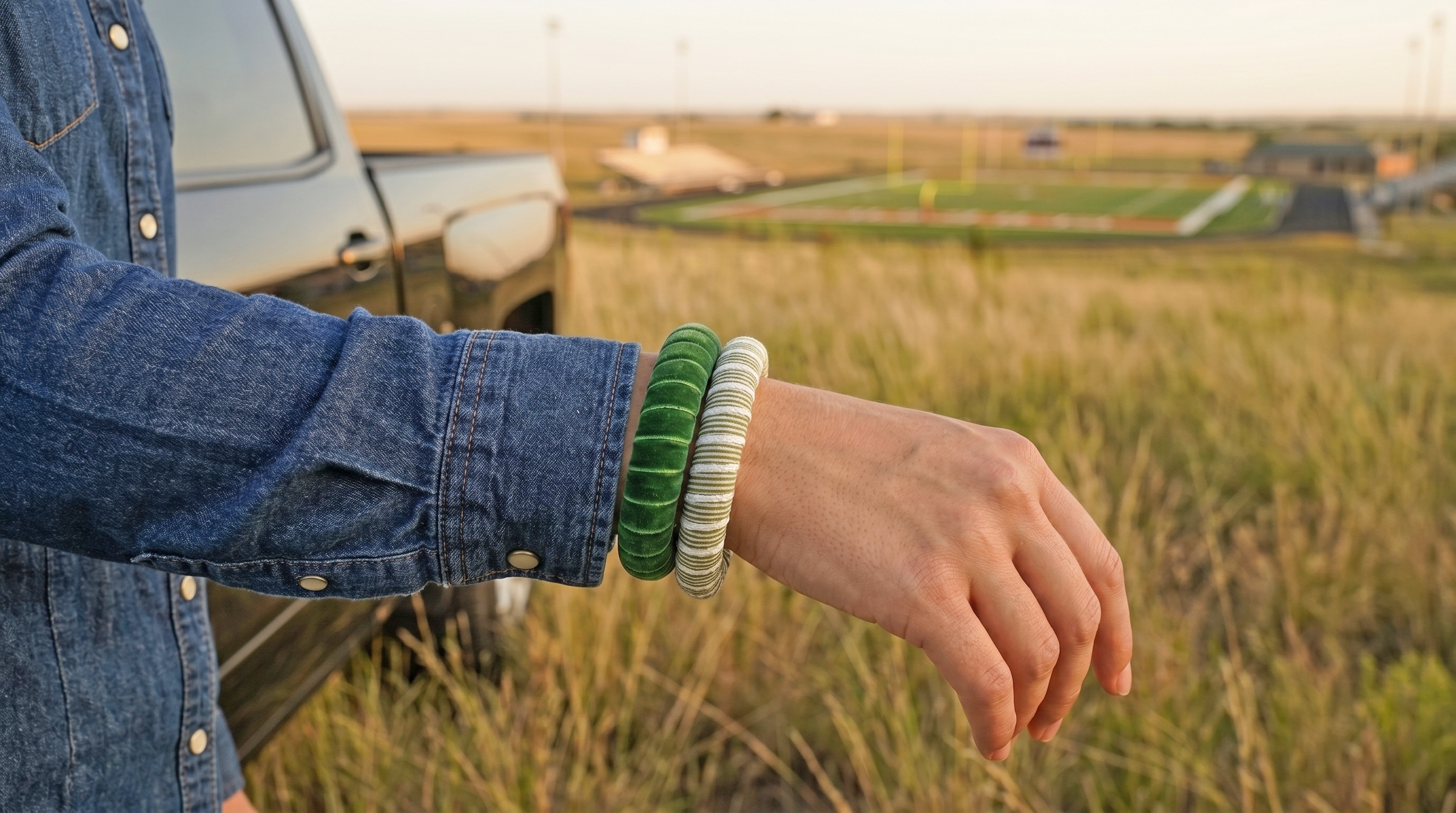 young woman wearing green and green and white Butter Republic bracelets and a denim shirt. She is in a grassy areas outdoors in a small town with a simple, small football field in the blurry background
