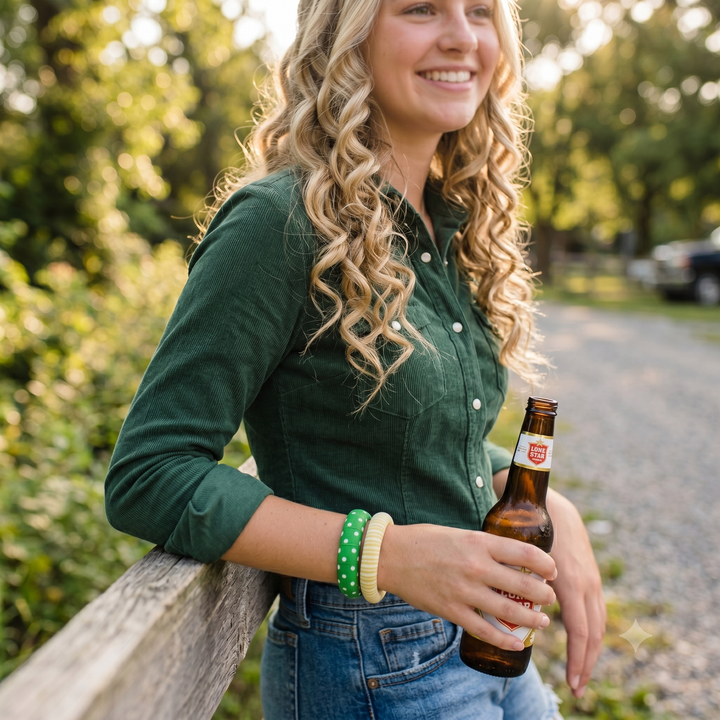 Young woman in a dark green shirt, wearing Butter Republic green and yellow bracelets, standing on the road drinking a beer out of a bottle