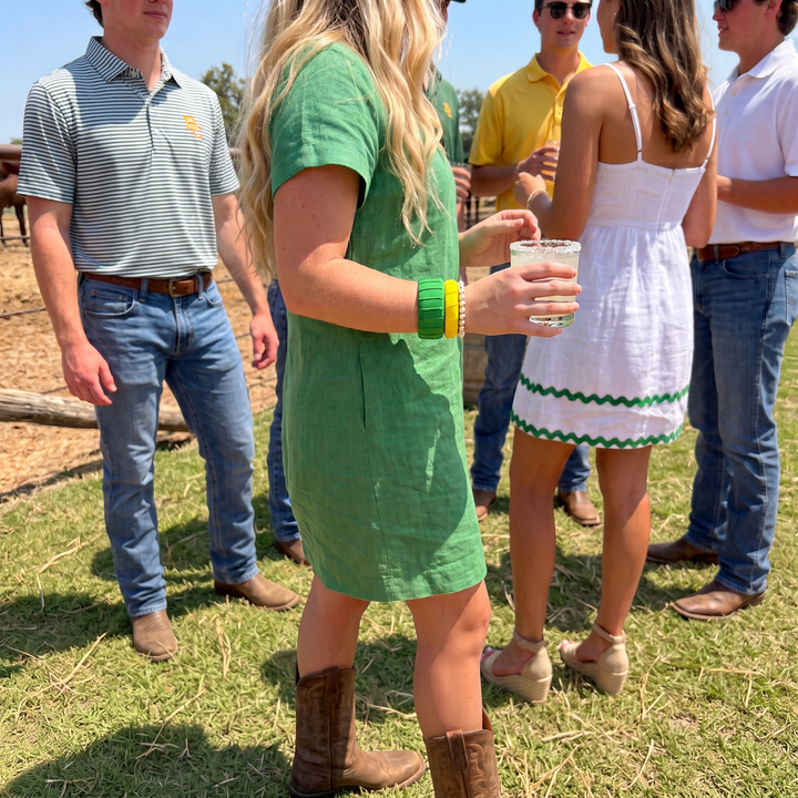 A woman in a green short-sleeve dress and brown cowboy boots, wearing a Butter Republic wide green bracelet and narrow yellow bracelet, stands outdoors at a sunny ranch or farm event, holding a drink. 