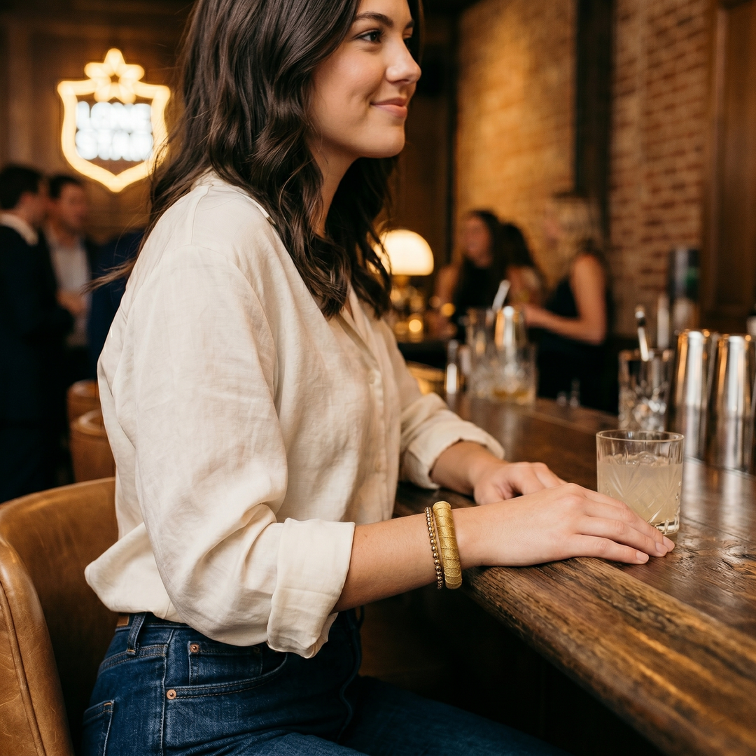 young woman wearing a gold Butter Republic bracelet, sitting at a bar, smiling