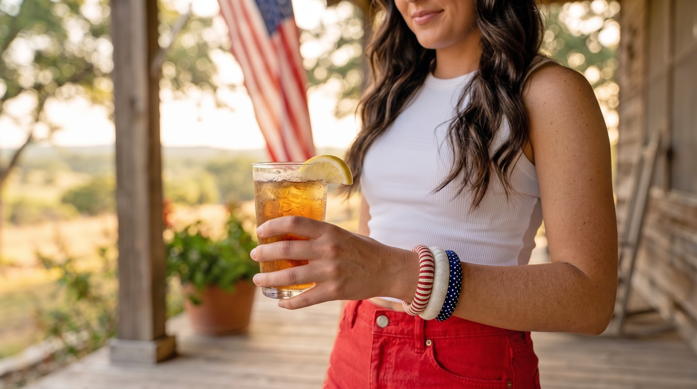 young woman wearing red, white, and blue Butter Republic bracelets, a white sleeveless top, and red denim shorts is standing on a wooden porch, holding a glass of iced tea with a blurred American flag waving in the background 
