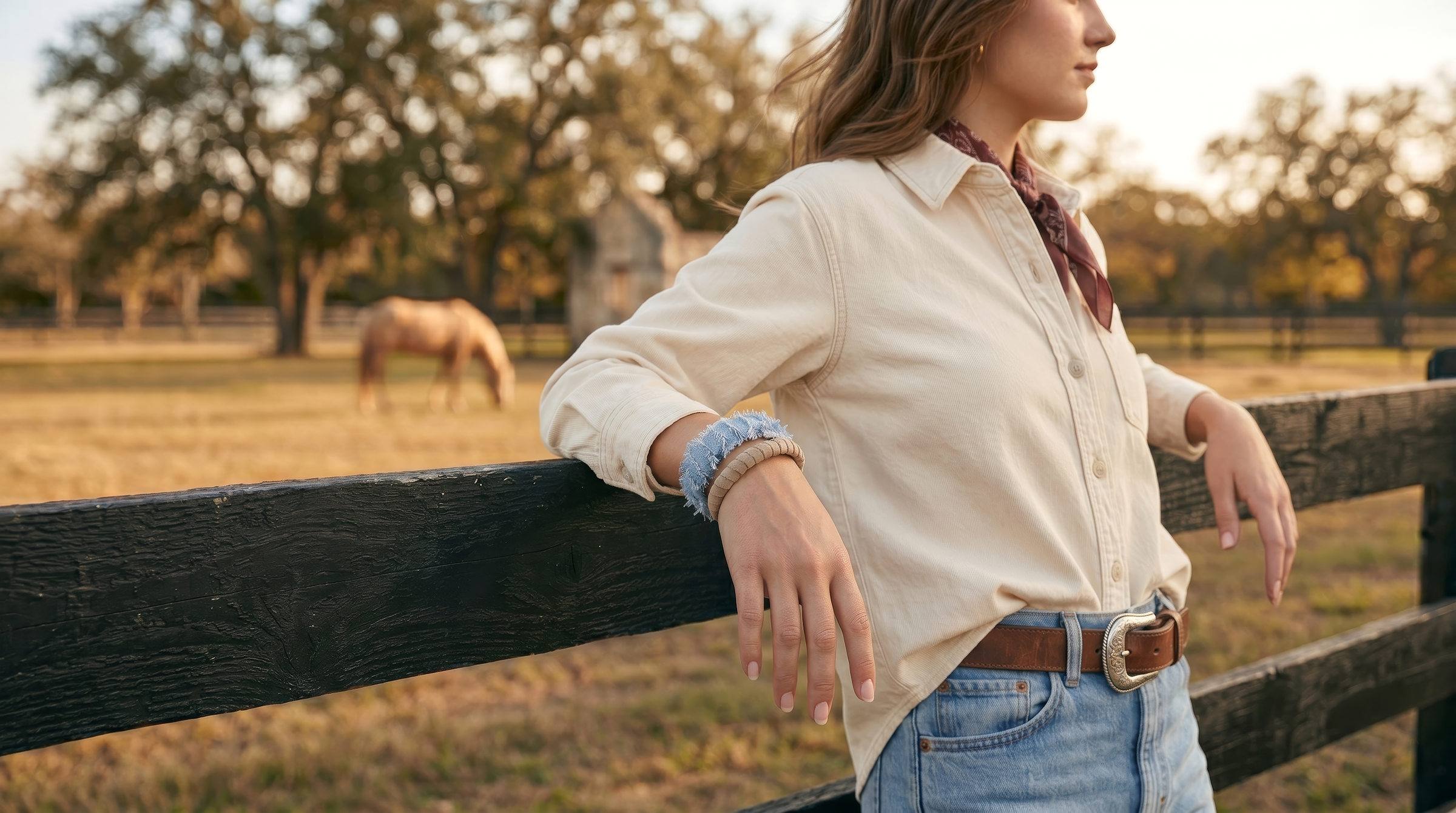 woman wearing light blue frayed and beige suede Butter Republic bracelets is standing by a wooden fence with a horse in the background on a farm