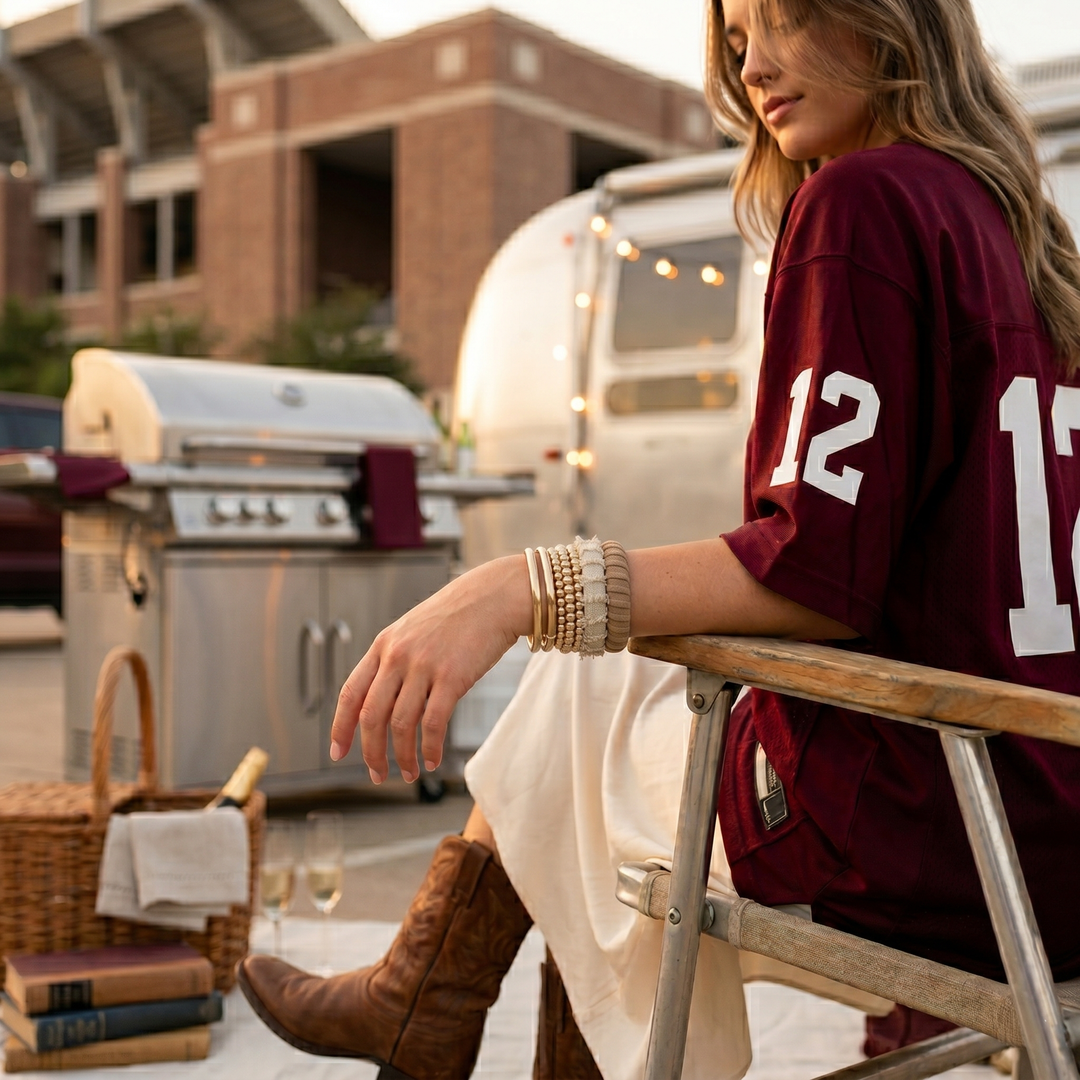young woman wearing two neutral colored Butter Republic bracelets and  a maroon and white jersey with number 12, sitting outdoors near a vintage camper
