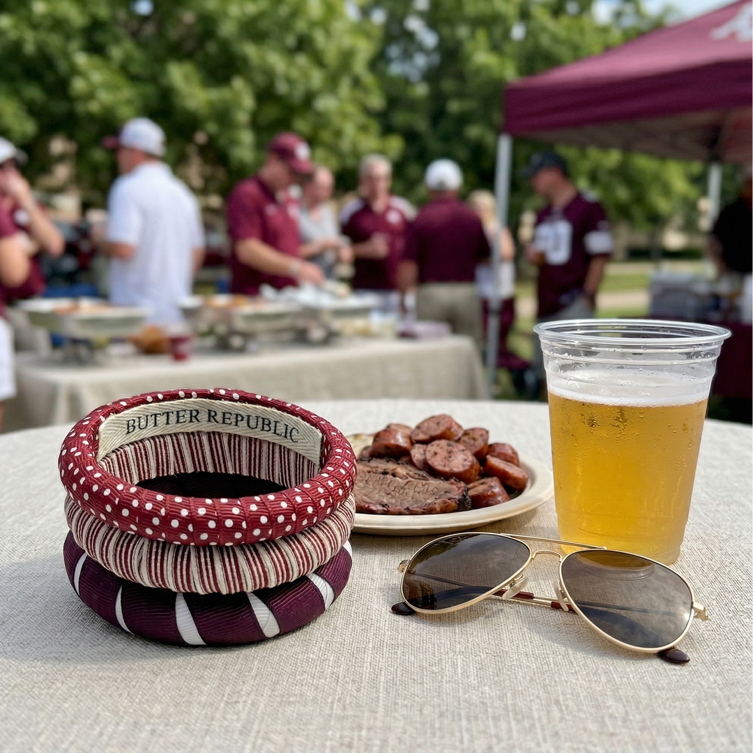 Butter Republic stack of maroon and white bracelets on an outdoor tailgate scene table with BBQ, sunglasses and beer.