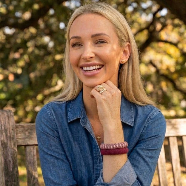 woman wearing two maroon Butter Republic bracelets, sitting outdoors on a wooden bench with blurry trees in the background