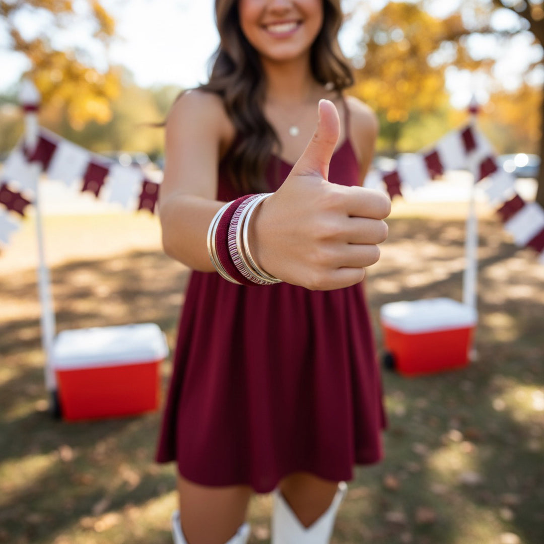 young woman in a maroon dress wearing maroon and white Butter Republic bracelets, giving a thumbs-up gesture outdoors with a blurred background of red coolers and maroon and white decoratve bunting
