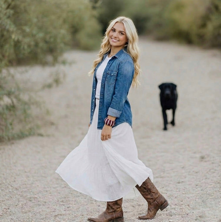 young woman in a white dress and blue denim shirt wearing a maroon and white Butter Republic bracelet,  walking on a dirt road with a black dog in the background