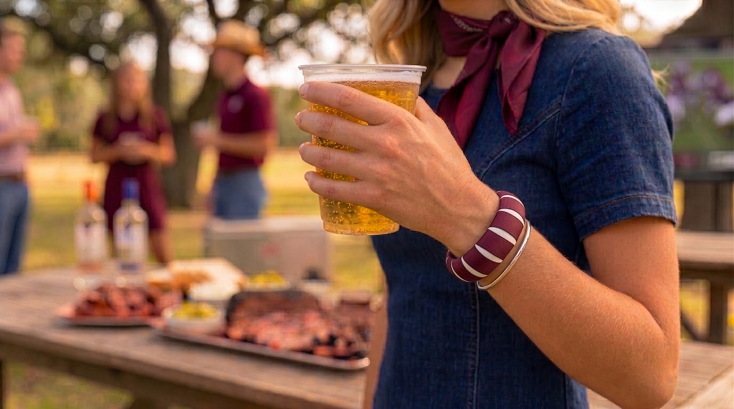 young woman outside, wearing a maroon and white Butter Republic bracelet and a dark blue denim dress, she is holding a cup of beer and standing in front of a table of BBQ