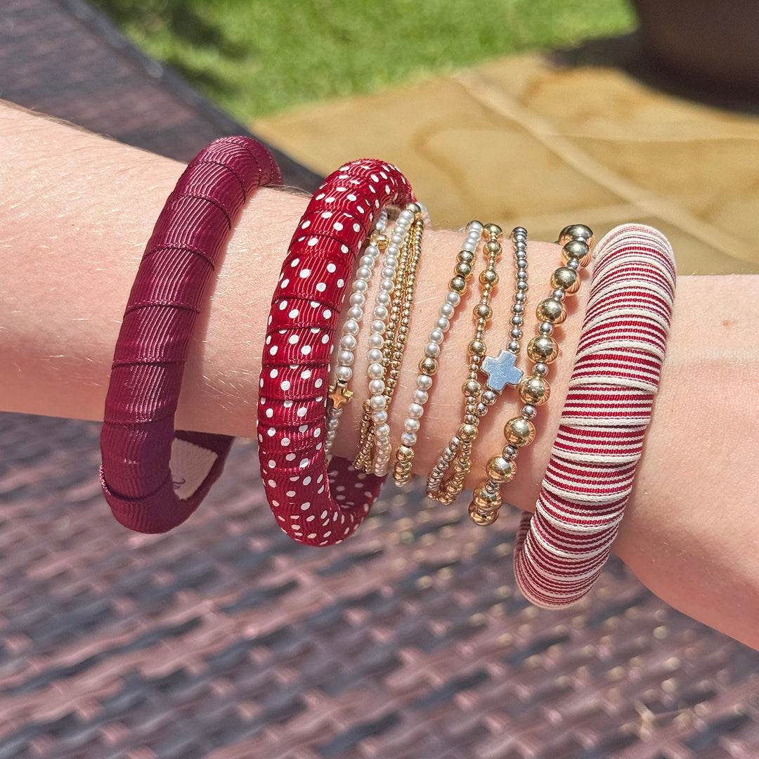 Close-up of a wrist wearing multiple maroon and white Butter Republic bracelets on a textured surface with grass in the background
