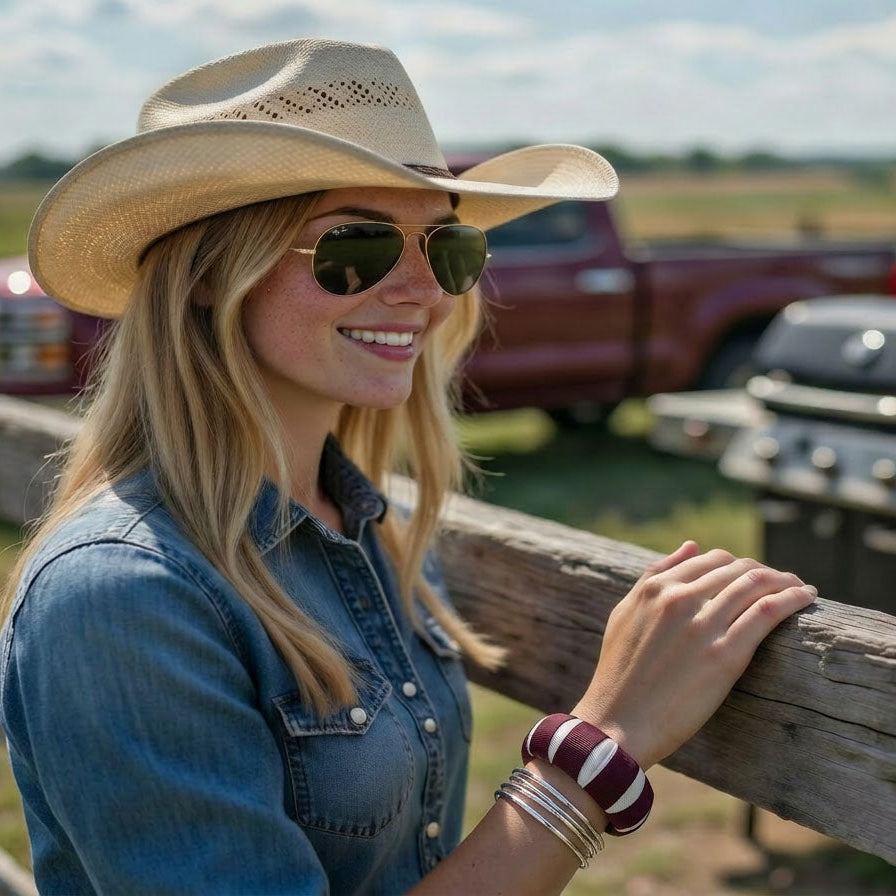 woman wearing a maroon and white Butter Republic bracelet, a cowboy hat, and aviator sunglasses, standing by a wooden fence with a maroon truck and grill in the background