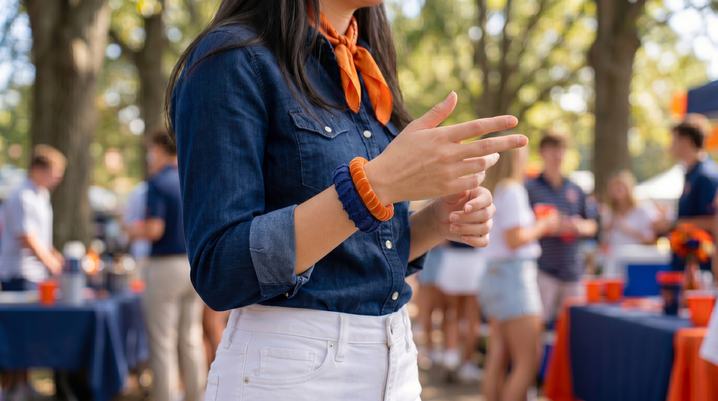 young woman at an outdoor tailgate gathering is wearing a frayed navy blue Butter Republic bracelet and a velvet orange Butter Republic bracelet, a denim shirt, white shorts, and an orange scarf around her neck. The background includes blurred fans and tables, gathered around tables