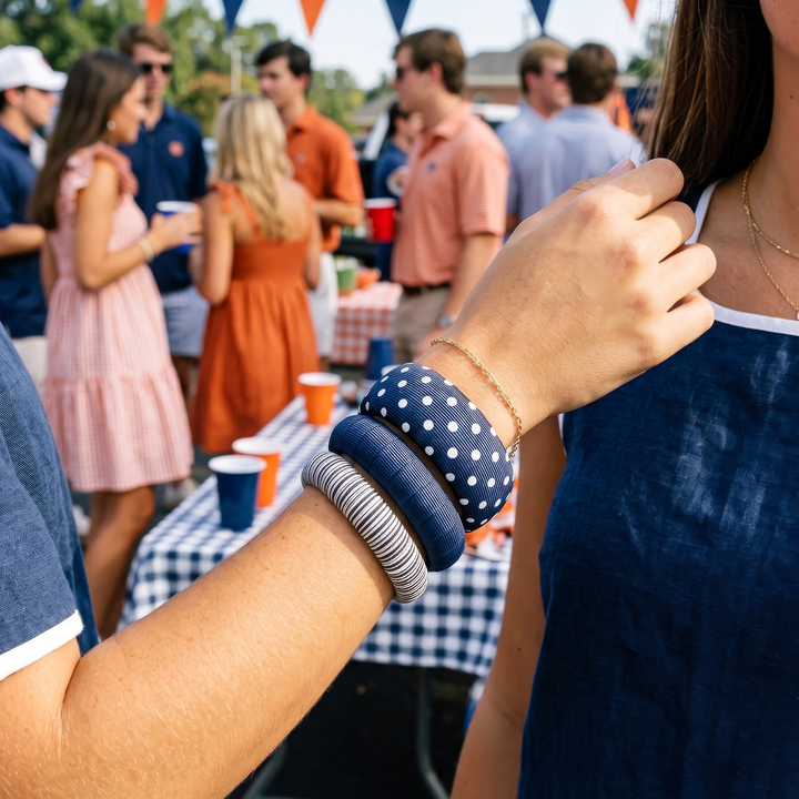 A woman wearing assorted navy blue and white Butter Republic bracelets at an outdoor tailgate event with a table and colorful orange and navy blue flags in the background