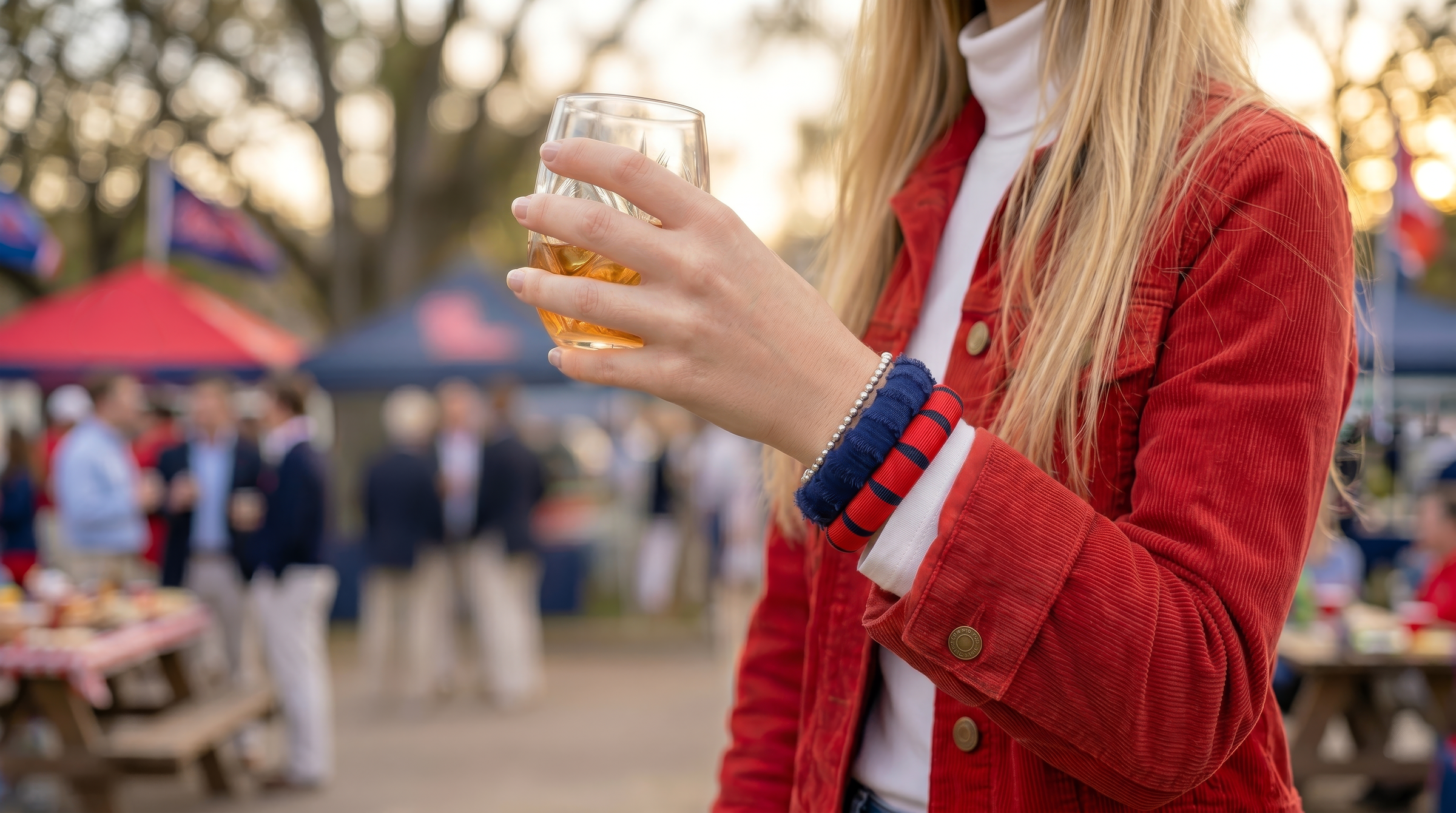young woman at an outdoor football tailgate in the fall, wearing a navy blue Butter Republic bracelet and a red and blue Butter Republic bracelet with a white turtleneck top nd a red corduroy jacket. Blurred background with multiple football fans, tailgate tents, picnic tables, and red and blue accent colors.