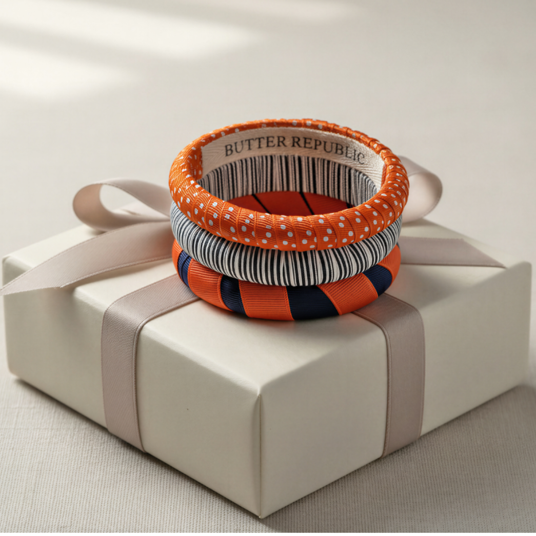 three orange and navy blue Butter Republic bracelets stacked on a beige ribbon-tied white box with Butter Republic logo brand label on the interior of the bracelet