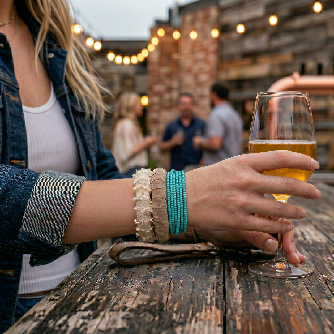 two neutral Butter Republic bracelts on the wrist of a woman seated at a wood table outside, holding a glass of beer at dusk with fairy lights blurry in the background