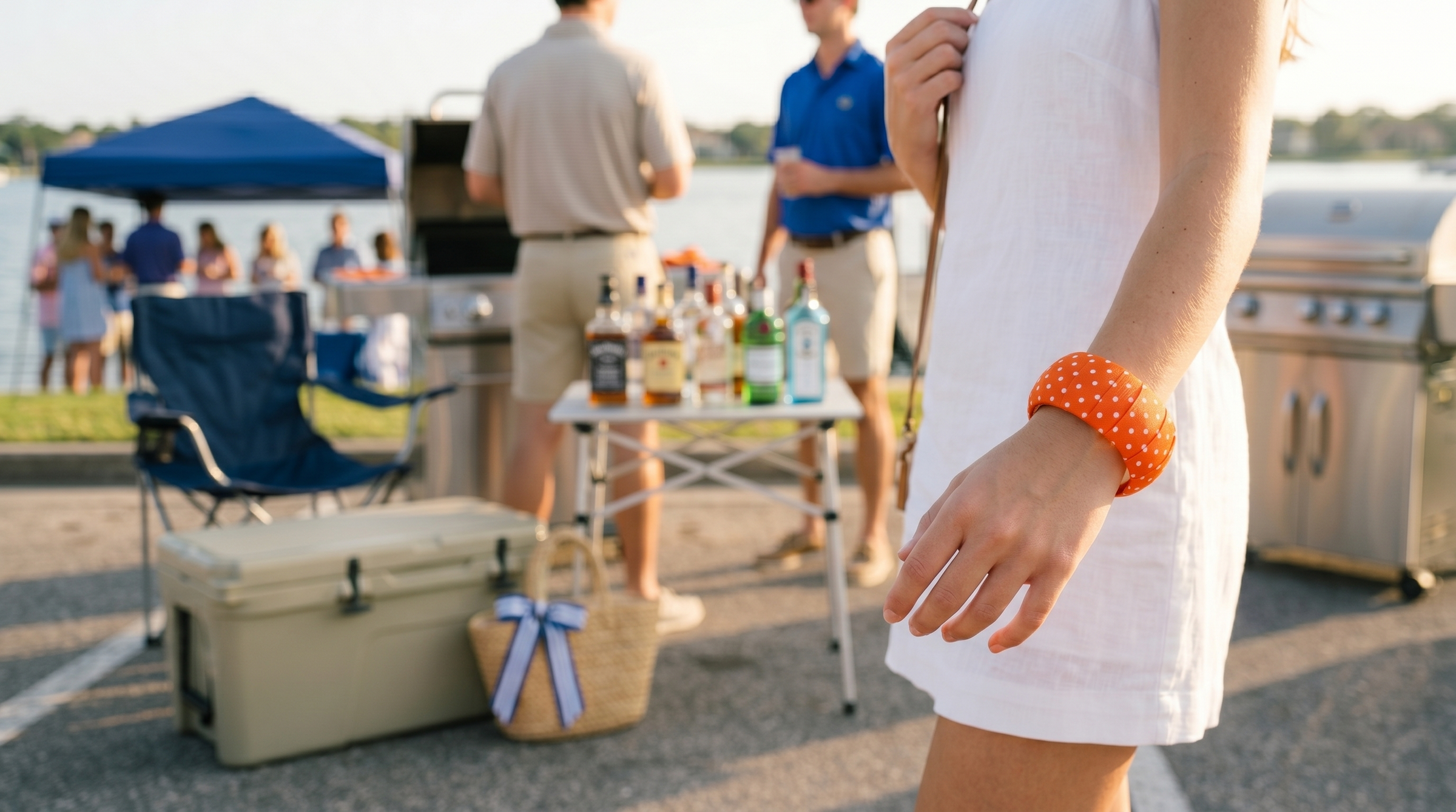 woman wearing an orange Butter Republic bracelet and a white shift dress is outdoors at a lake party with blurry guests in the background behind her