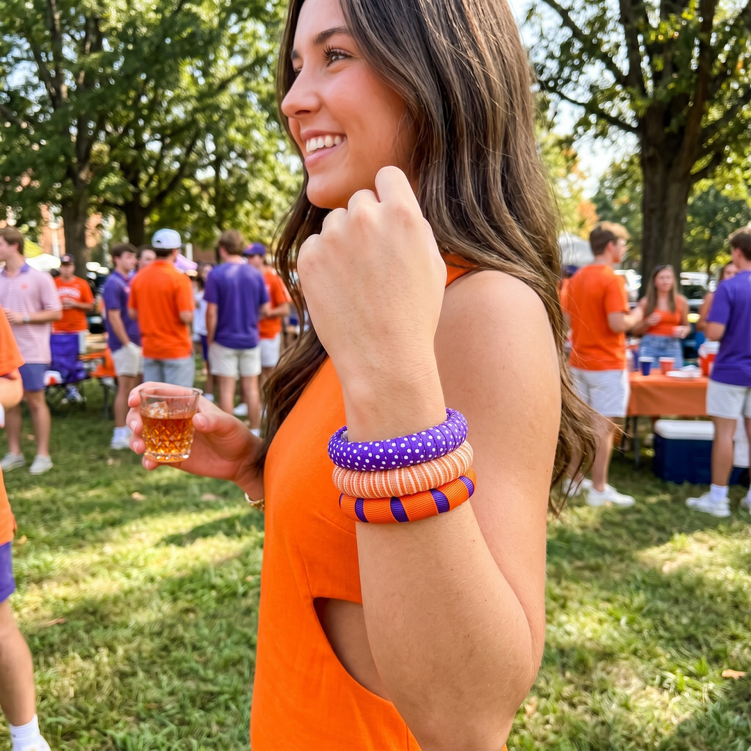 Woman wearing an assortment of orange, purple, and white Butter Republic bangles, in an orange dress holding a drink, surrounded by people in a casual outdoor setting.