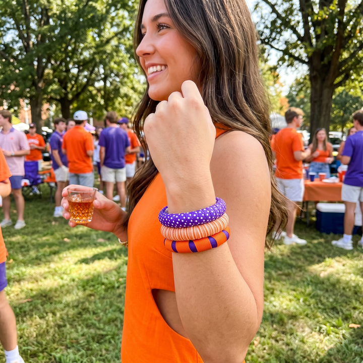 Woman wearing an assortment of orange, purple, and white Butter Republic bangles, in an orange dress holding a drink, surrounded by people in a casual outdoor setting.