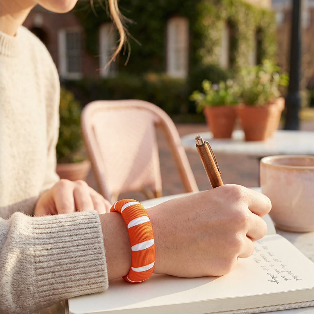woman holding a pen and wearing a Butter Republic orange and white bracelet, sitting outdoors with a notebook and cup