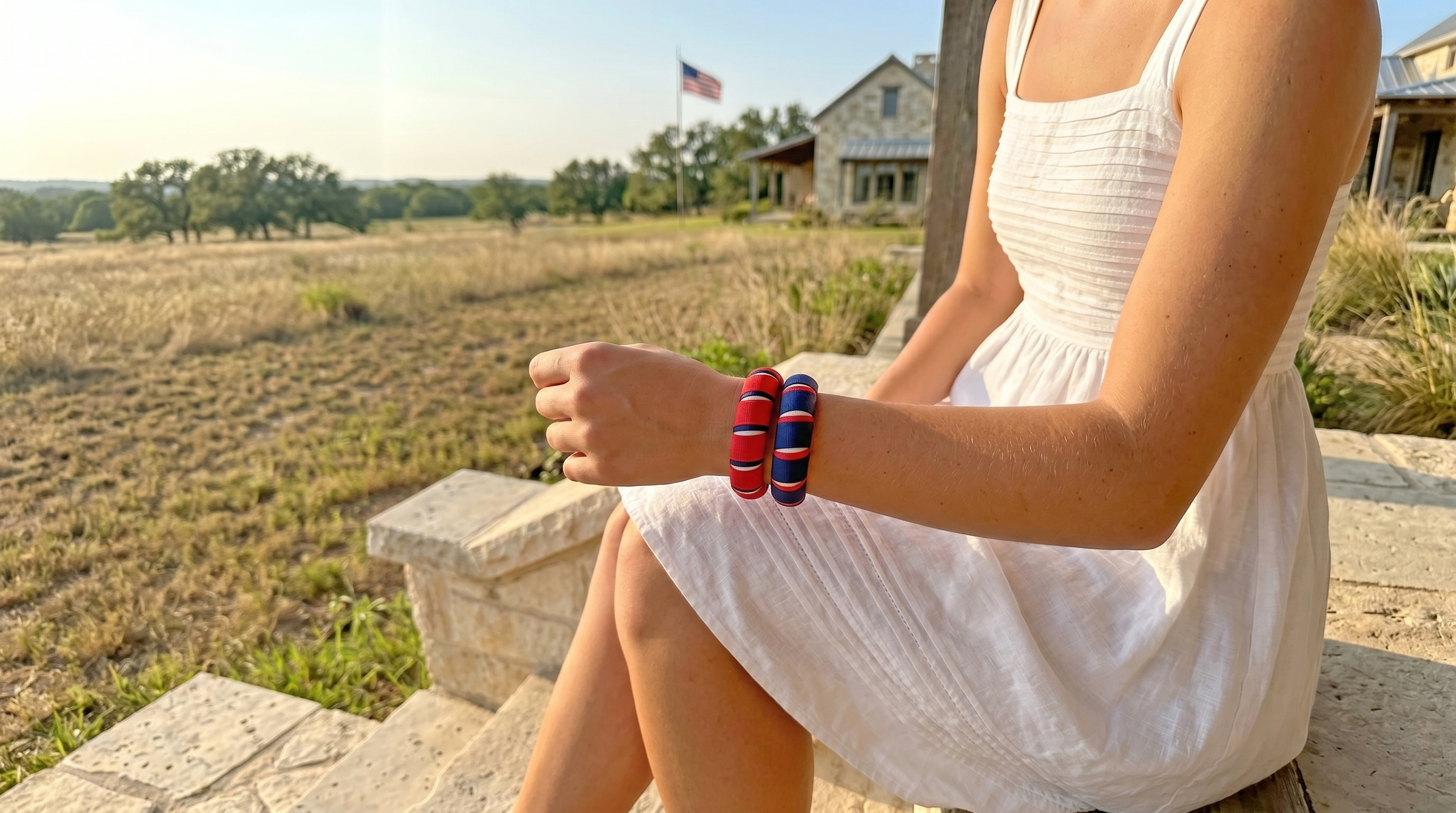 woman wearing two red, white, and blue Butter Republic bracelets and a white dress, sitting outdoors with a scenic background