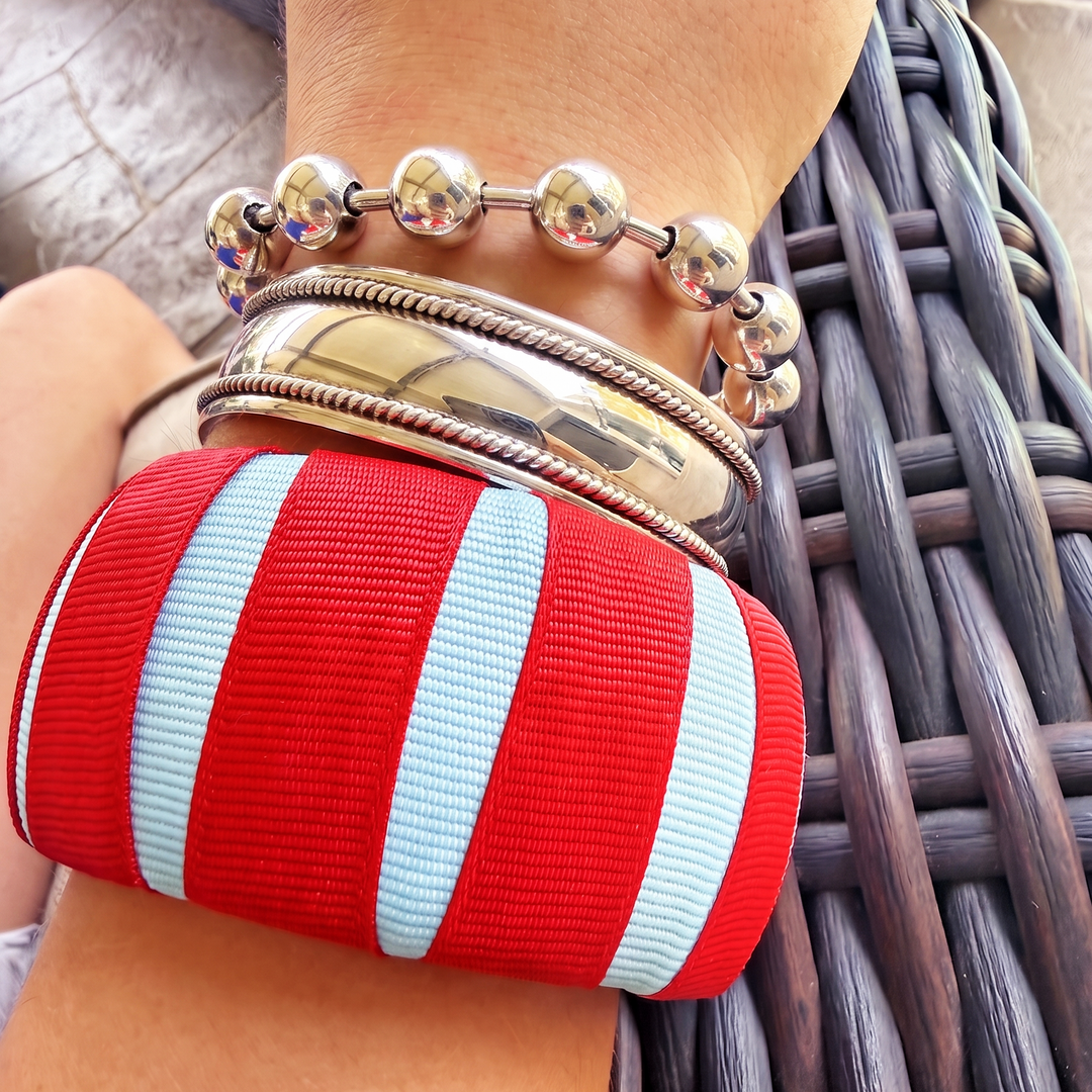 Close-up of a wrist wearing a powder blue and red Butter Republic bracelet with multiple silver bracelets on a woven surface
