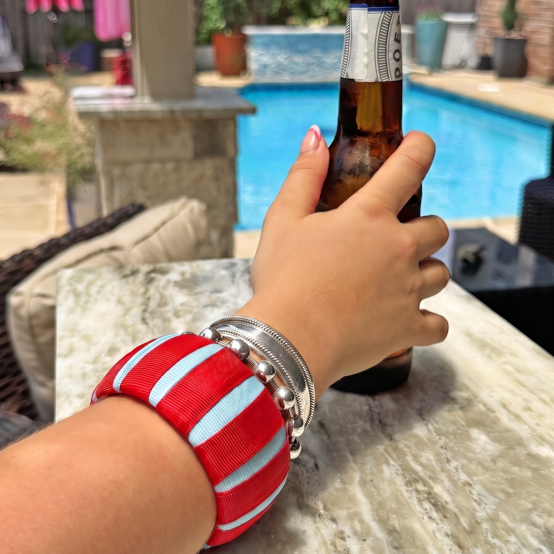 Hand, wearing a Butter Republic bracelet, holding a beer bottle with a pool and outdoor furniture in the background