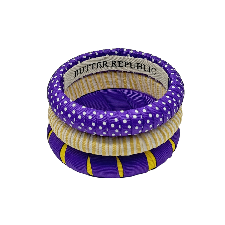 a stack of three Butter Republic bracelets in purple and white, yellow and white, and purple and gold, with a logo brand label on the interior of the bracelets