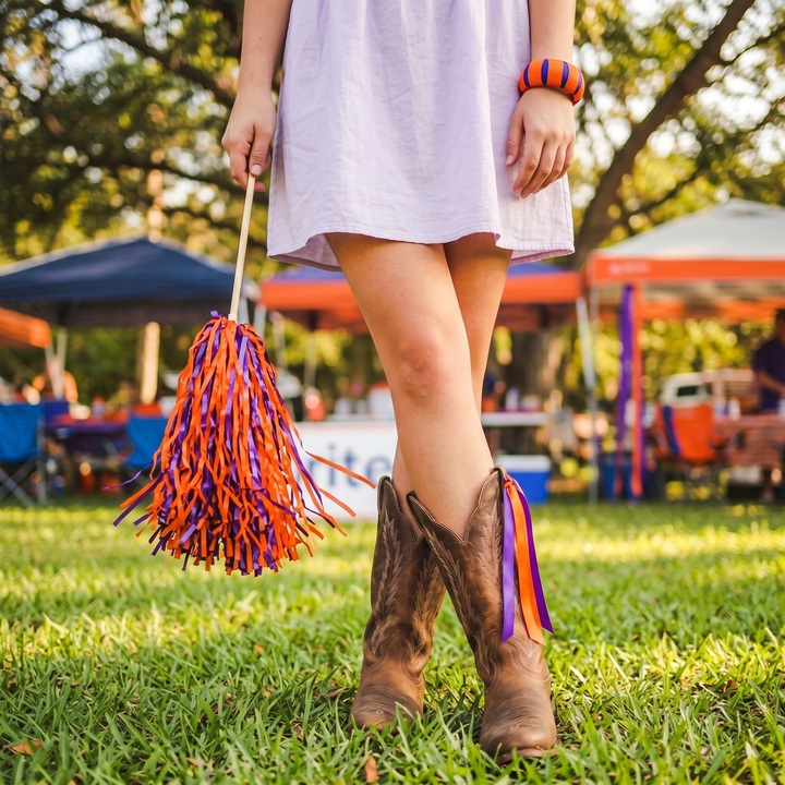 A girl wearing a purple and orange Butter Republic bracelet, holding an orange and purple pom-pom in a grassy outdoor setting with tents and people in the background.