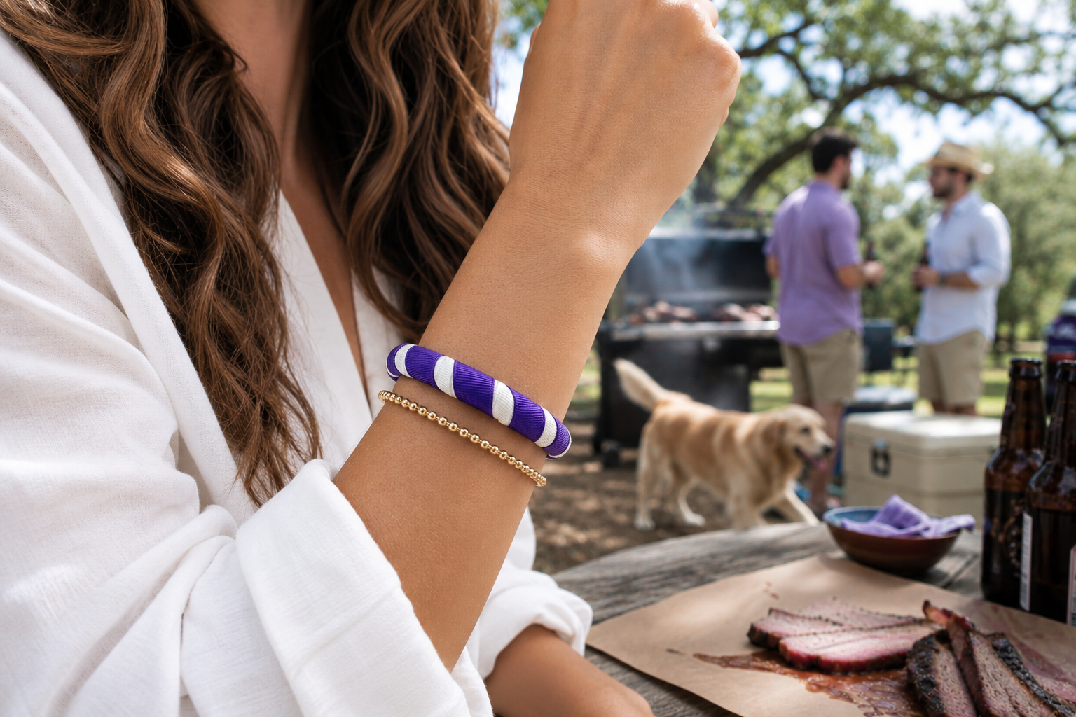 woman is wearing a purple and white Butter Republic bracelet outdoors with a blurred view behind her of a dog and two gents talking in front of a grill