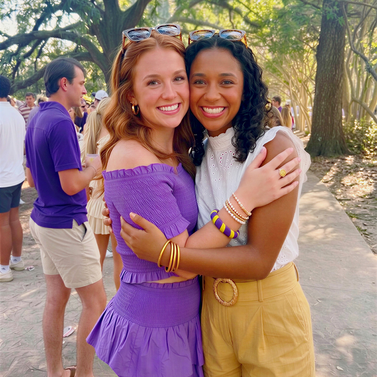 two young women, one wearing a purple and yellow Butter Republic bracelet, hugging outdoors with trees and people in the background