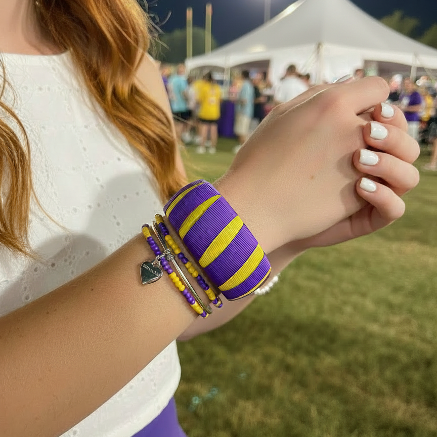 woman wearing a purple and yellow Butter Republic bracelet with multiple purple and gold beaded bracelets in a grassy tailgate area at night
