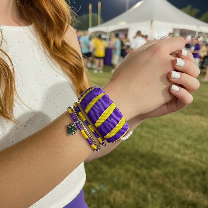 woman wearing a purple and yellow Butter Republic bracelet with multiple purple and gold beaded bracelets in a grassy tailgate area at night