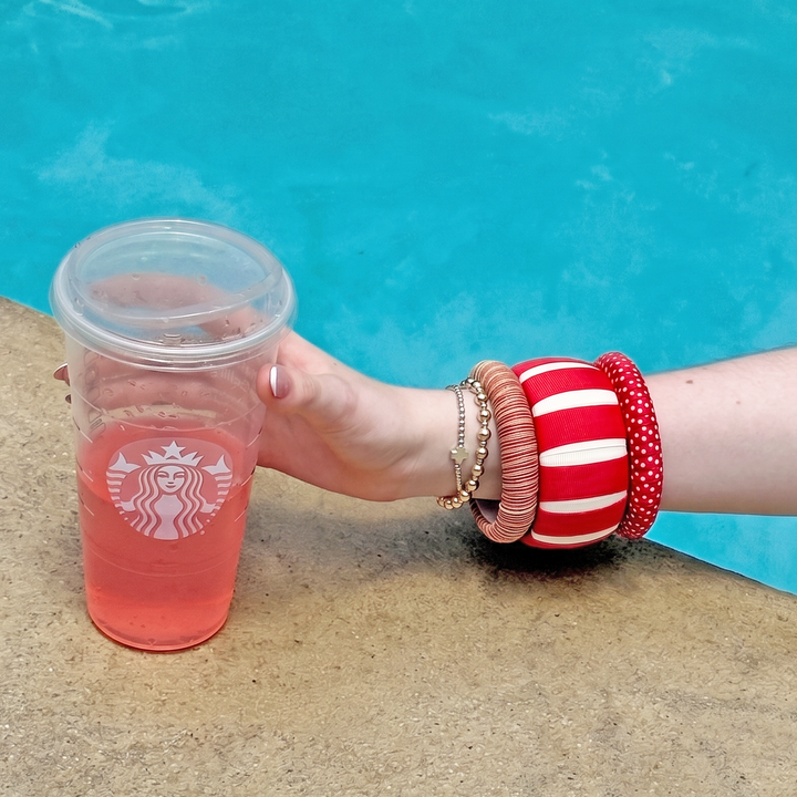 Hand wearing red and white Butter Republic bracelets, holding a pink Starbucks cup by a pool