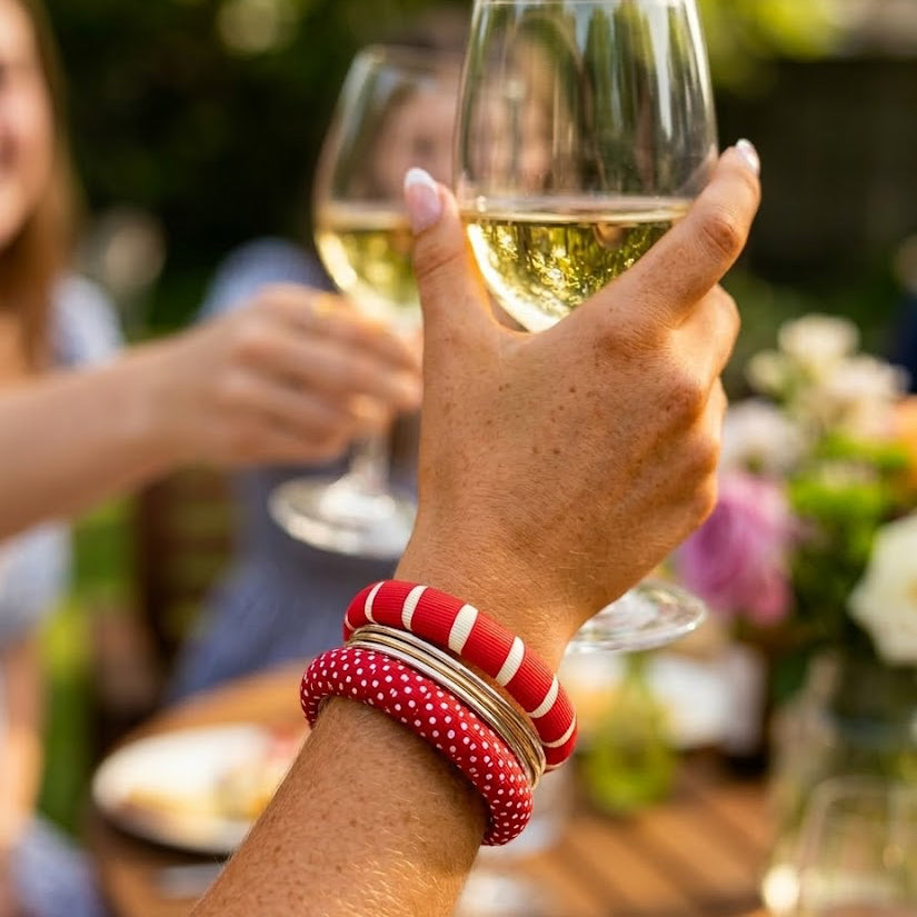 woman holding a glass of white wine with a blurred outdoor setting, wearing two red and white Butter Republic bangle bracelets