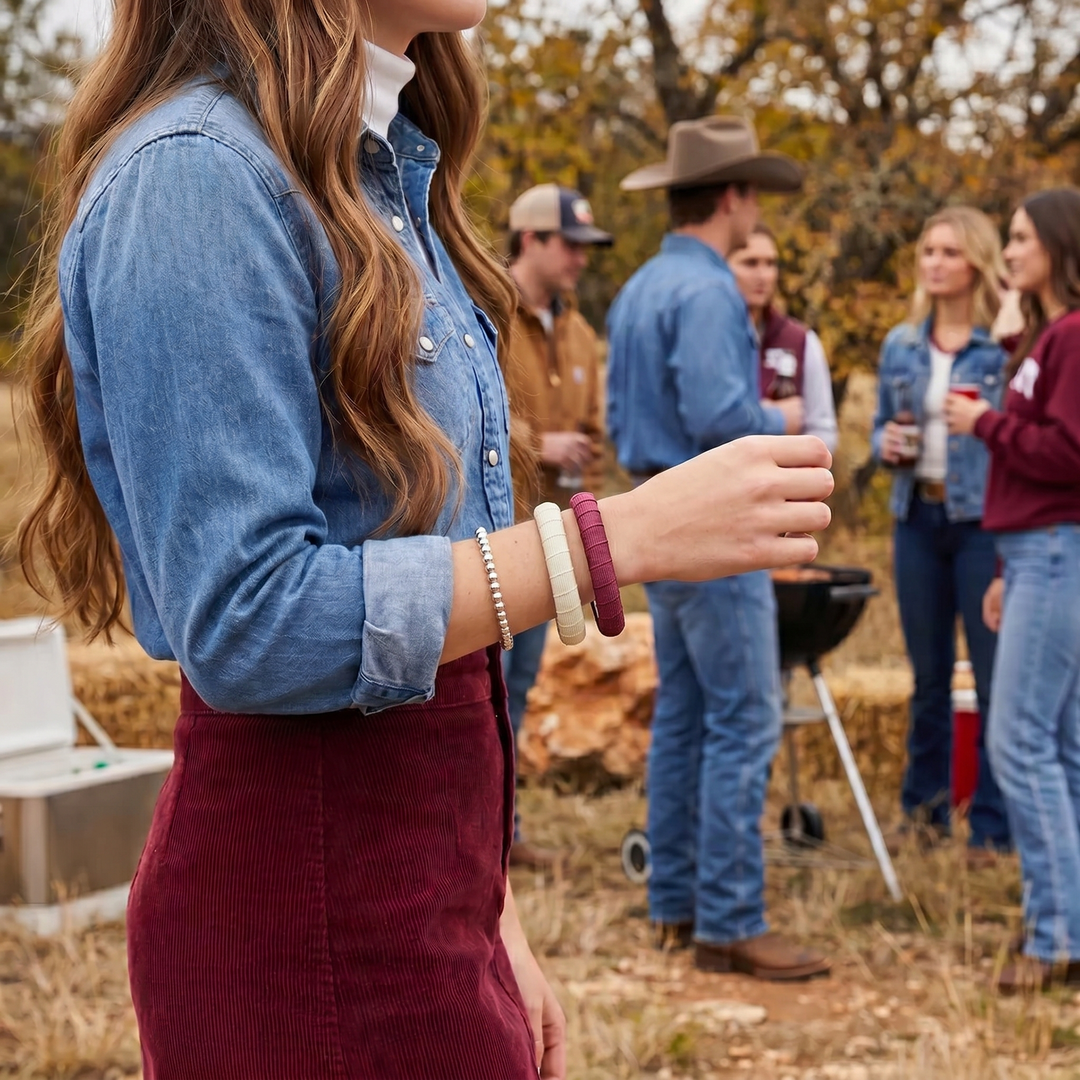woman wearing a maroon Butter Republic bracelet and a cream Butter Republic bracelet and a blue shirt and maroon skirt with a group of people standing around a small black grill in a field