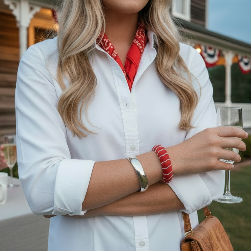 woman in a white shirt with rolled-up sleeves, wearing a red and white moonstitch Butter Republic bracelet, holding a glass of champagne, standing outdoors near a table with a cup and plate