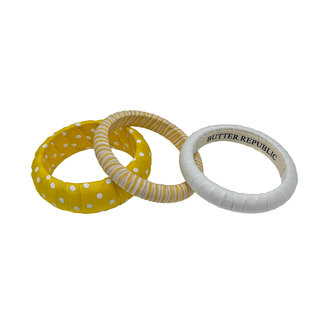 Three yellow and white Butter Republic bracelets in polka dots, stripes, and solid white; brand label is affixed to the interior 