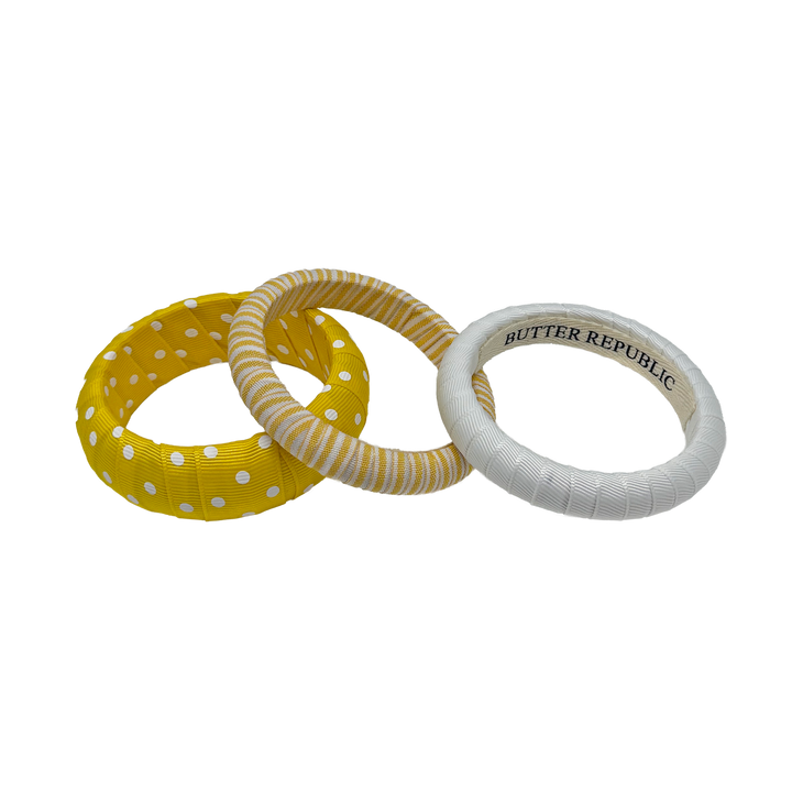 Three yellow and white Butter Republic bracelets in polka dots, stripes, and solid white; brand label is affixed to the interior 